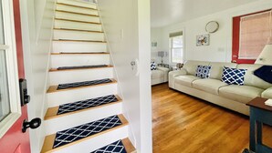 Una escalera con peldaños de madera y un corredor estampado que lleva a una sala de estar con un sofá color crema, cojines estampados y una pequeña mesa.