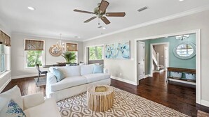 Ein geräumiges Wohnzimmer mit weißem Sofa, einem gemusterten Teppich, Deckenventilator und Kunstwerken an den Wänden.