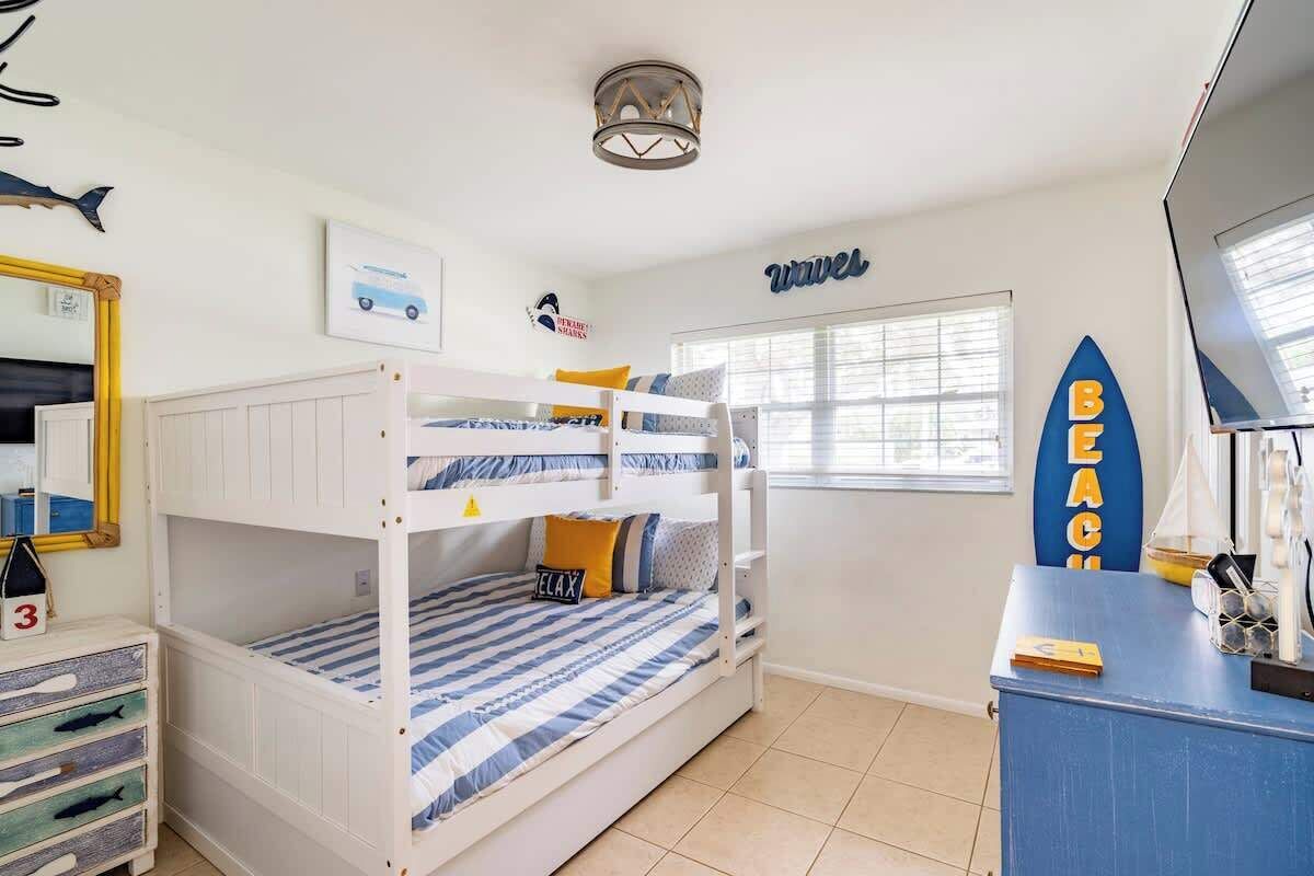 Een slaapkamer met stapelbed, een decoratie van een surfplank, een bureau met computer en een raam met jaloezieën.