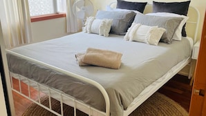 Un lit bien fait, avec une couette grise et blanche, un plaid blanc et plusieurs coussins aux couleurs gris, blanc et noir. Un tapis rond en fibres naturelles est placé sous le lit, et un ventilateur blanc se trouve sur le sol, près de la fenêtre.