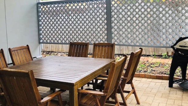 Un espace extérieur aménagé en terrasse, avec une table et des chaises en bois, des guirlandes lumineuses suspendues et une vue sur la verdure à travers une clôture en treillis.