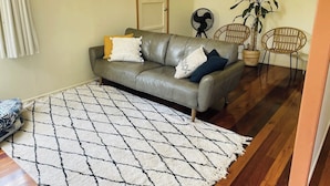 Un salon avec un canapé en cuir gris, un tapis à motifs, un ventilateur et deux fauteuils en osier.