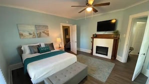 A bedroom with a bed, a bench, a fireplace, a ceiling fan, and a TV.