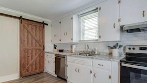 A kitchen with white cabinets, a wooden sliding door, a dishwasher, a stove, and a window with blinds.