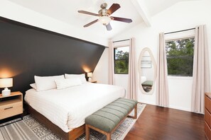 Une chambre à coucher comprenant un lit, des tables de chevet, un banc, un ventilateur de plafond et une fenêtre avec des rideaux.