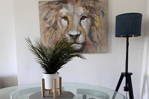 A room with a glass table, a potted plant, a painting of a lion's face, and a floor lamp.