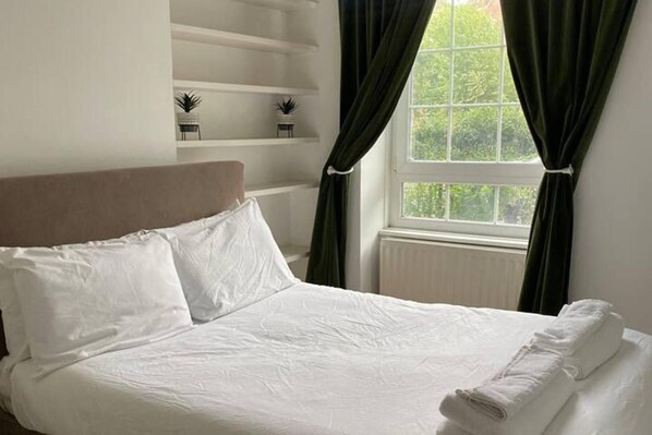 A neatly made bed with white linens, a headboard, and a window with curtains.
