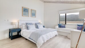 A bedroom with a bed, bedside tables, a bench by the window, and framed artwork on the wall. - Bolinas Beach Home - Over the water with spectacular views (Bolinas)