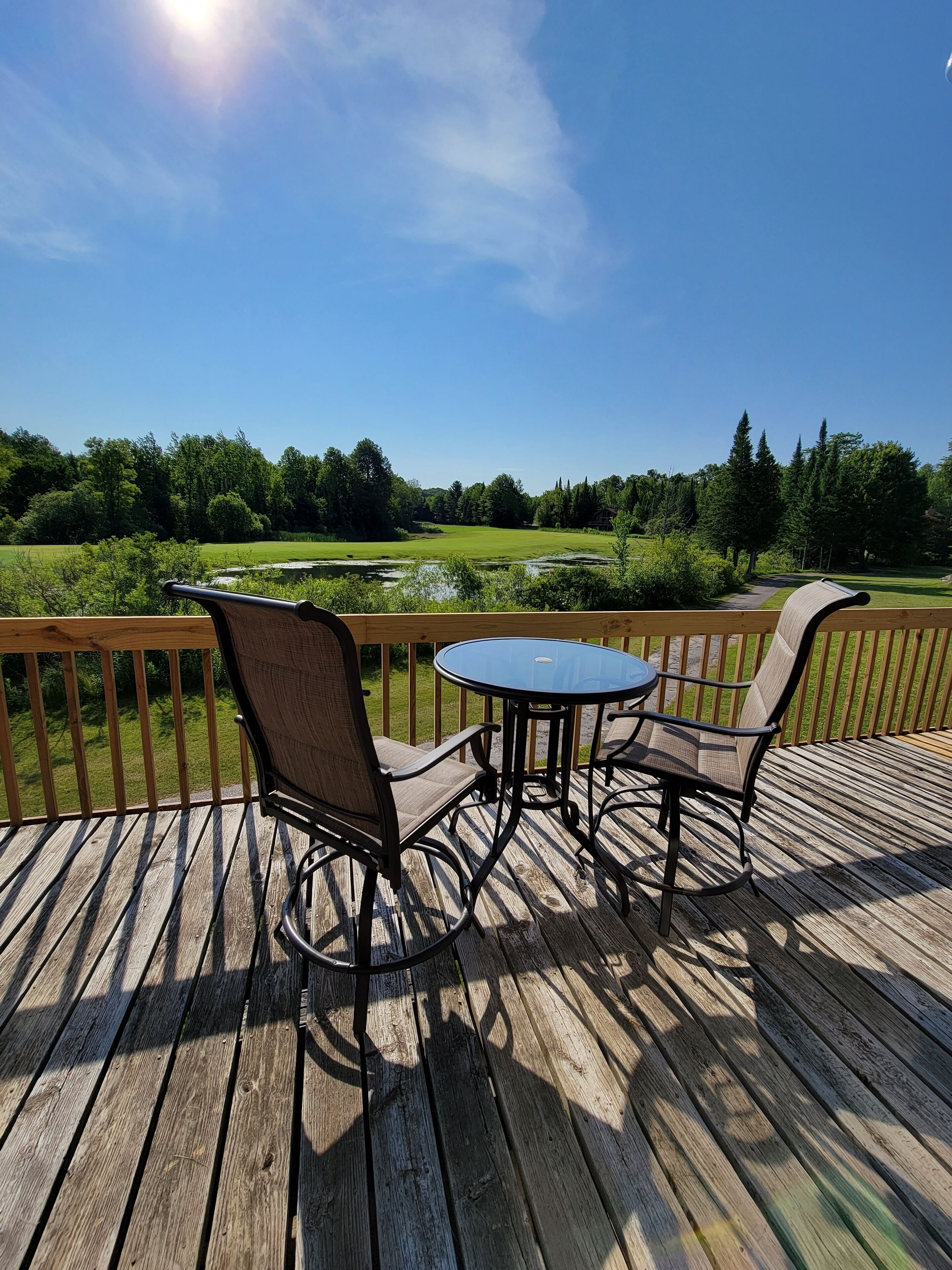 A wooden deck with a round table and two chairs, overlooking a grassy area with trees and a pond.