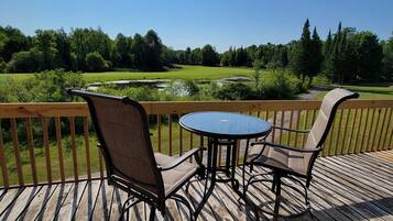 A wooden deck with a round table and two chairs, overlooking a grassy area with trees and a pond.