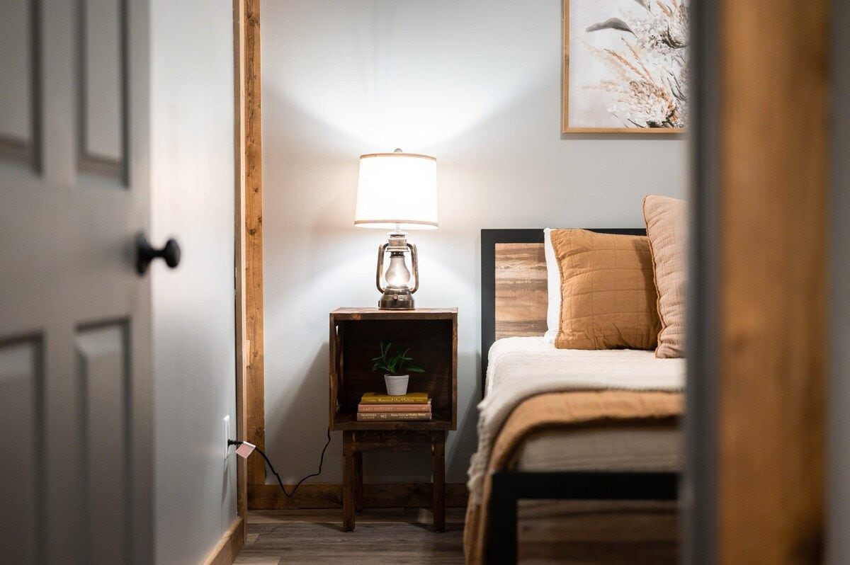 A bedroom with a bed, a bedside table with a lamp, a small shelf with books and a plant, and a wall-mounted painting.