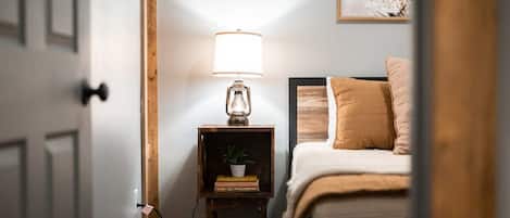 A bedroom with a bed, a bedside table with a lamp, a small shelf with books and a plant, and a wall-mounted painting.
