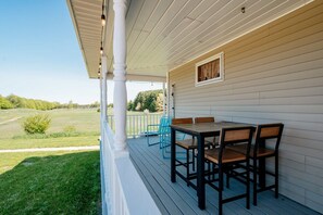 Eine überdachte Veranda mit Tisch und Stühlen, Blick auf ein saftiges Grasfeld und ein Fenster mit Vorhängen.