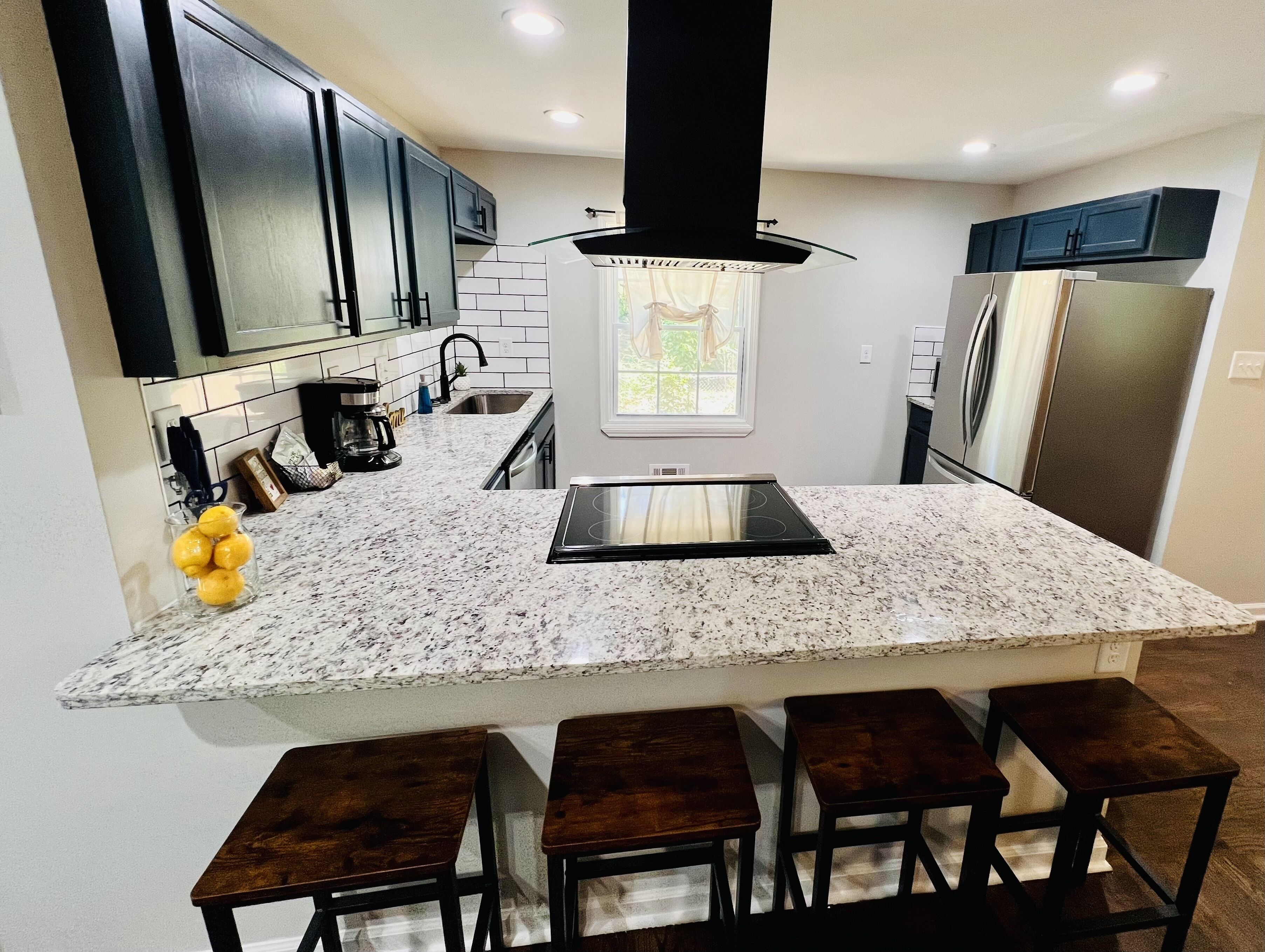 A kitchen with a central island, dark cabinets, a stainless steel refrigerator, and a window with curtains.