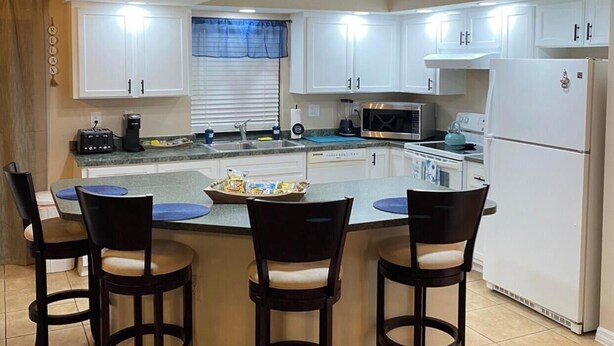 A kitchen with white cabinets, a central island with bar stools, and decorative items on the walls. - Cozy, Clean, Convenient With A Beach Feel (Cape Coral)