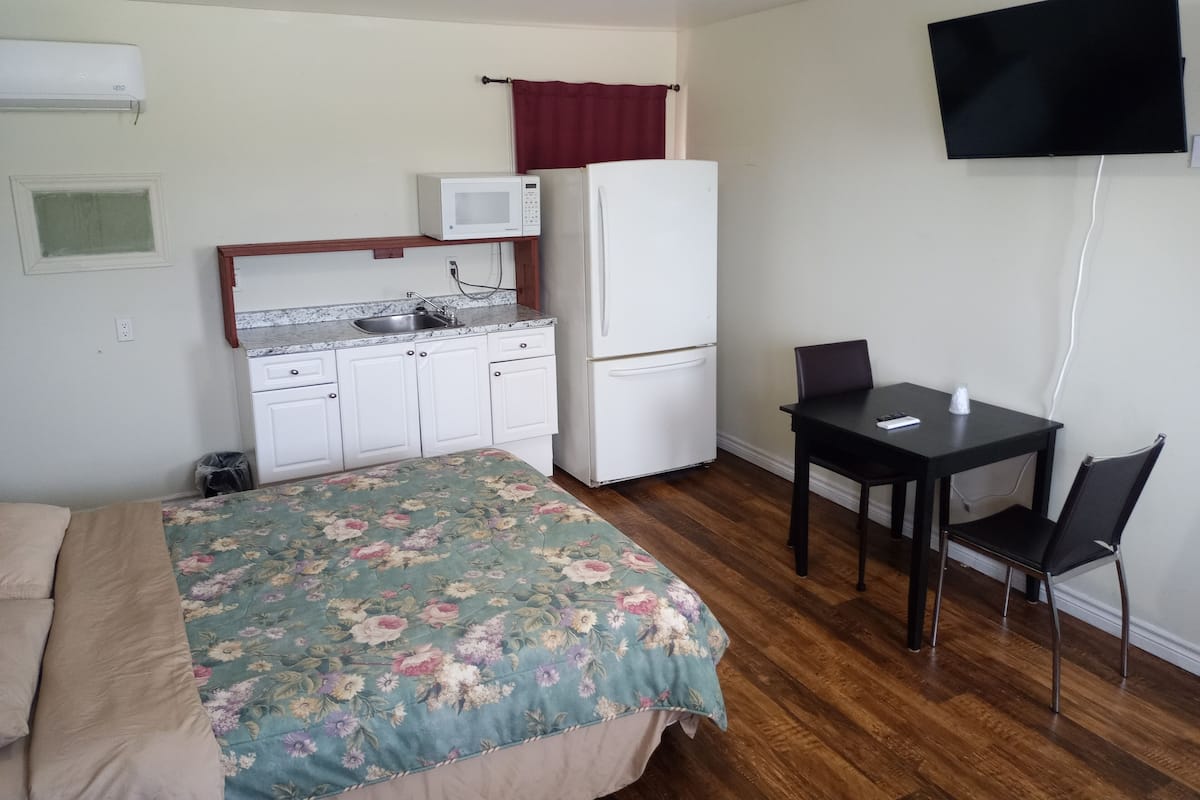 Una habitación compacta con cama, una kitchenette pequeña, televisor y un escritorio con silla.