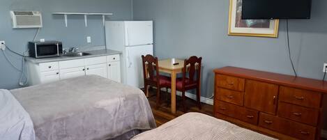 A room with two beds, a small kitchenette with a microwave and refrigerator, a wooden dresser, a dining table with chairs, and a TV mounted on the wall.