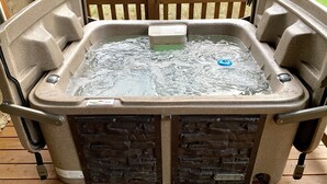 A hot tub with a stone facade, a control panel, and a view of a grassy field through a wooden deck. - Black Bear Retreat • 2Bed 2Bath Cabin • Peaceful Retreat (Townsend)