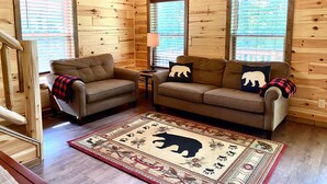 A cozy cabin interior with wooden walls, two sofas with animal-themed cushions, a patterned rug, and a staircase. - Black Bear Retreat • 2Bed 2Bath Cabin • Peaceful Retreat (Townsend)