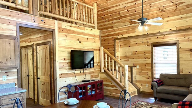 A wooden interior with a dining area, a television mounted on the wall, a staircase, and a sofa. - Black Bear Retreat • 2Bed 2Bath Cabin • Peaceful Retreat (Townsend)
