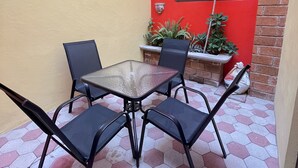 Un petit espace patio extérieur avec une table à plateau en verre et quatre chaises, entouré d’un mur jaune et d’un mur rouge agrémenté d’une fenêtre et de guirlandes lumineuses décoratives.