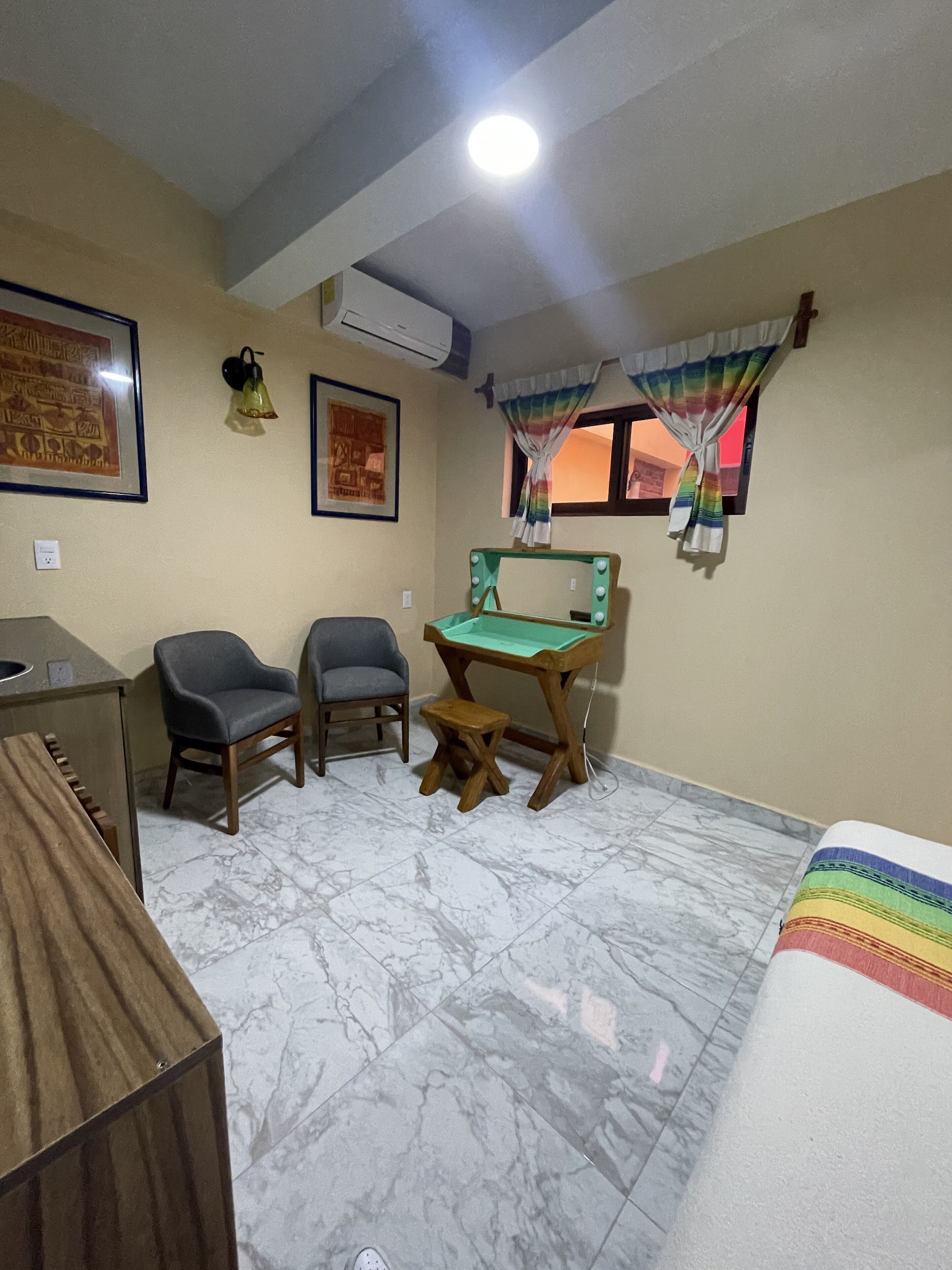 A room with a desk, two chairs, a small stool, a bed, a wall-mounted air conditioner, a framed picture, and a window with curtains.