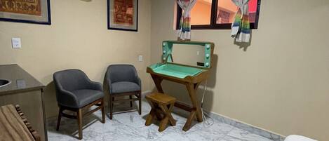 A room with a desk, two chairs, a small stool, a bed, a wall-mounted air conditioner, a framed picture, and a window with curtains.
