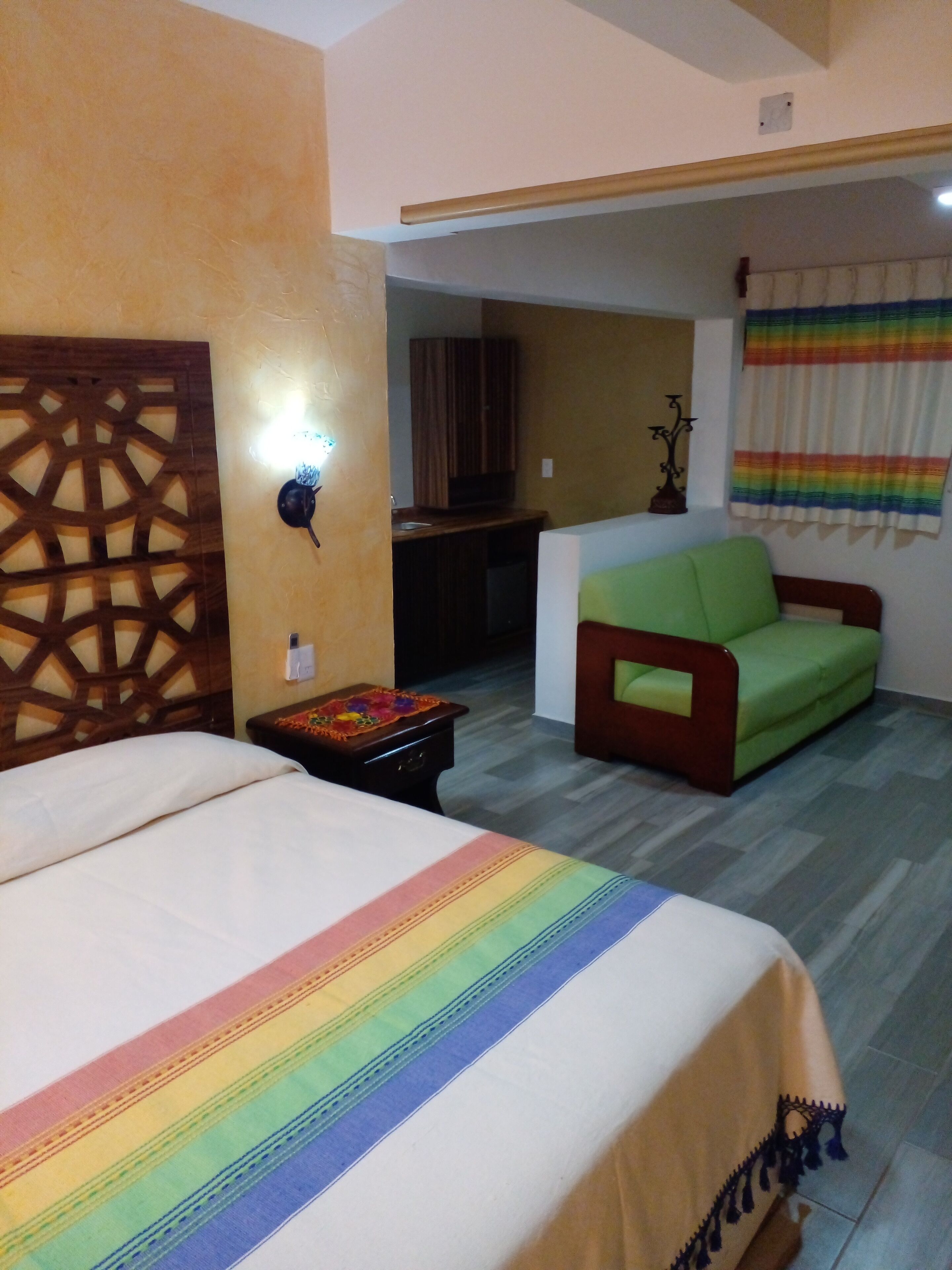 A hotel room with a bed, a green sofa, a small table with a decorative item, and a wall-mounted light fixture.