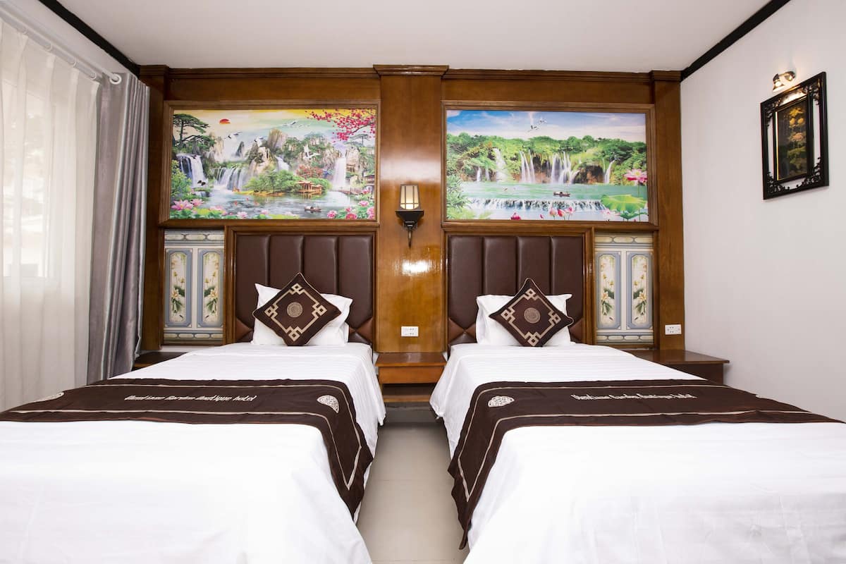 A hotel room with two beds, each with a headboard and pillow. There are two wall-mounted light fixtures and a framed picture above the beds.
