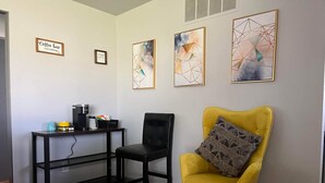 Una habitación con un sillón amarillo, una silla negra, una estación de café y tres cuadros abstractos en la pared.