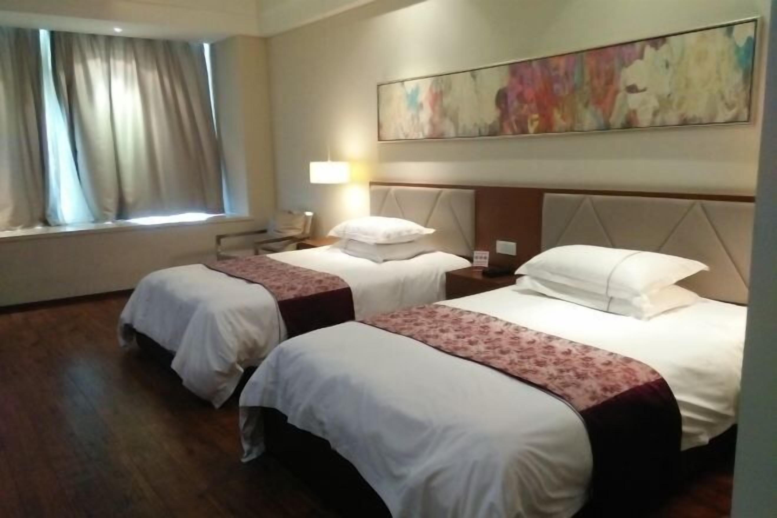 A hotel room with two beds, a large window with curtains, a wall-mounted painting, and a bedside table with a lamp.