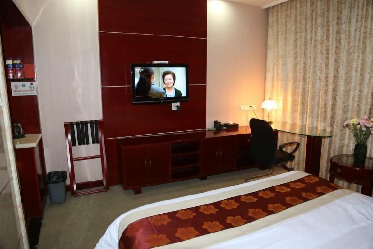Habitació d'hotel amb un llit, un televisor muntat a la paret, escriptori, cadira, làmpada i una taula petita amb un vas.