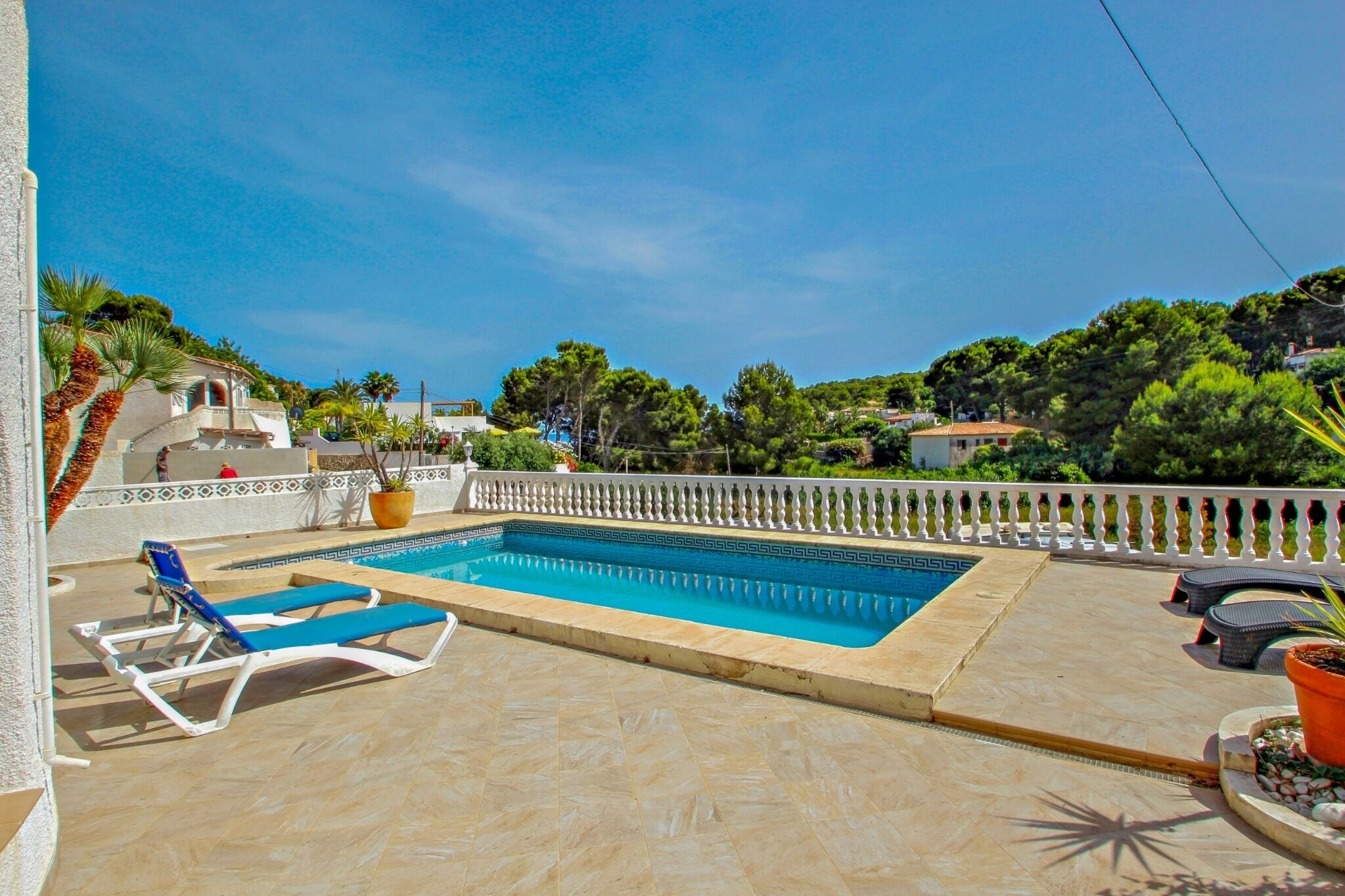 A rectangular swimming pool with blue water, surrounded by a white balustrade. There are two blue lounge chairs and several potted plants.