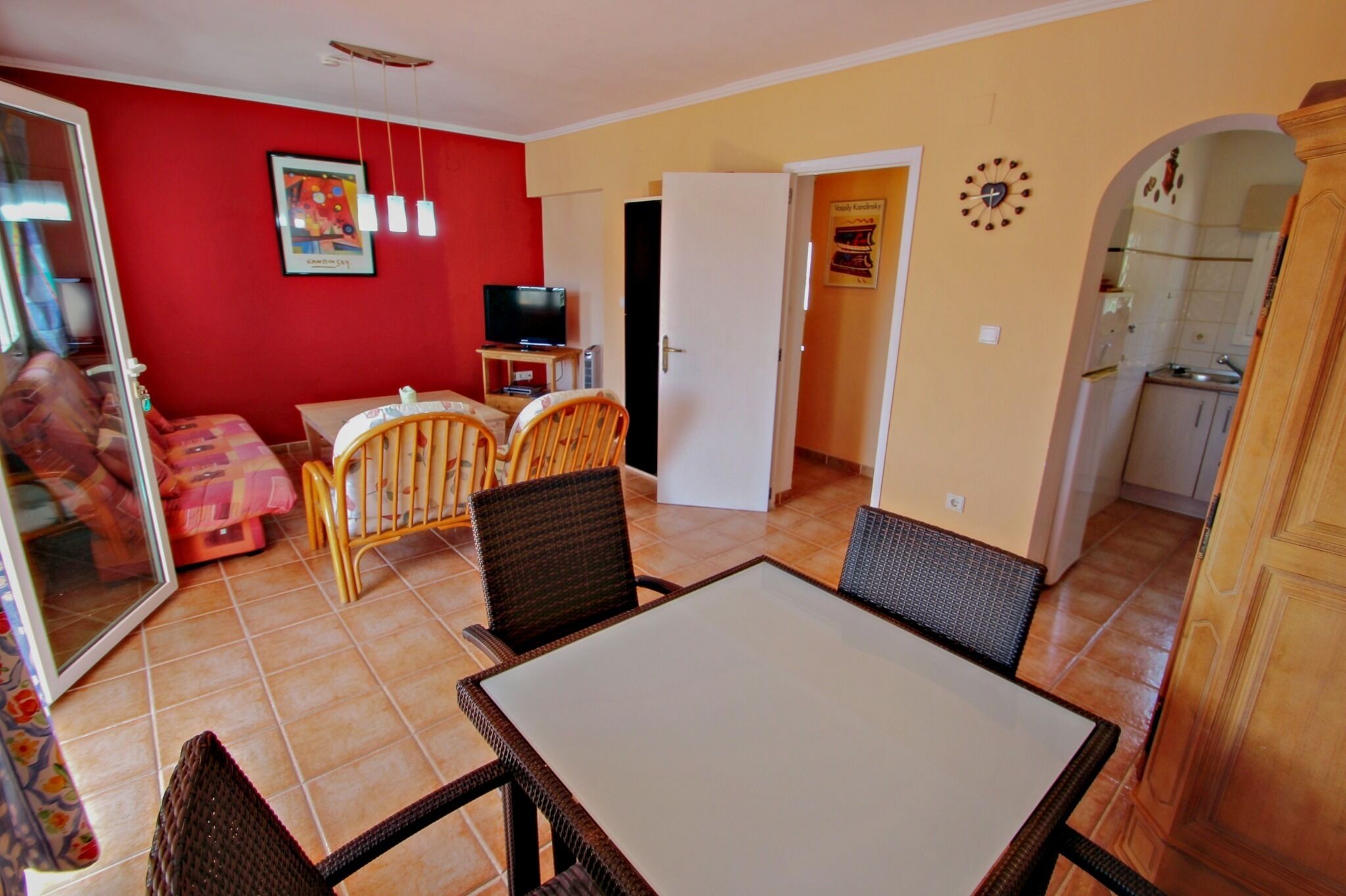 Un coin repas avec une table et des chaises, un téléviseur posé sur un meuble, et un mur rouge orné d’une affiche.