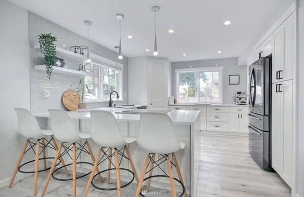 Una cocina moderna con armarios blancos, una isla central con taburetes y luminarias colgantes, además de un gran ventanal que proporciona luz natural.