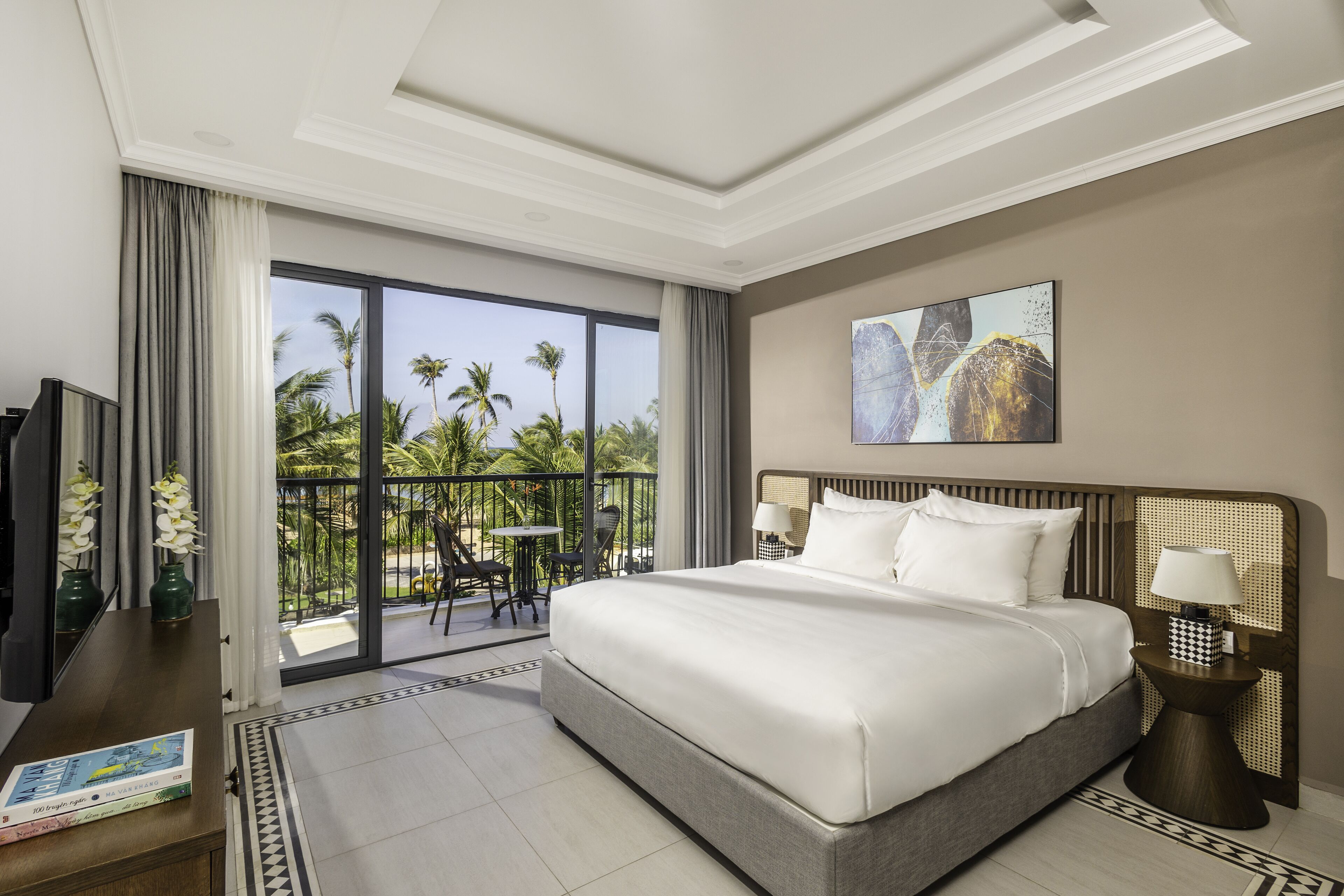 A hotel room with a large bed, a TV, a balcony with a view of palm trees, and a painting on the wall.