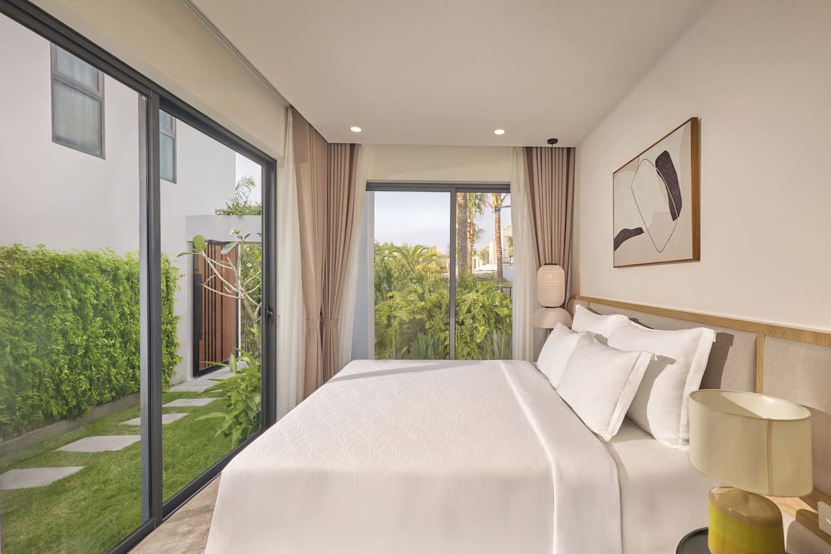 A hotel room with a large bed, a view of greenery, and a painting on the wall.