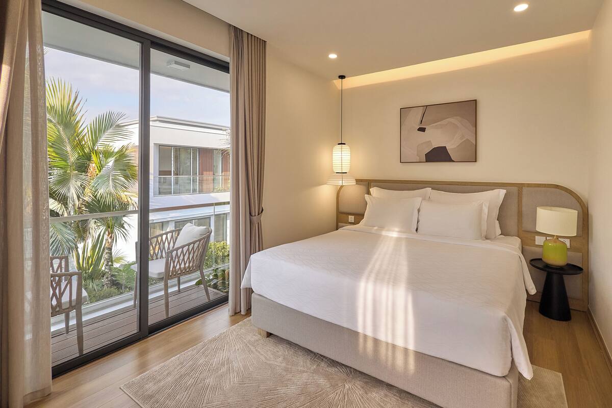 A hotel room with a bed, a nightstand, a lamp, a painting, and a view of palm trees through a sliding door.