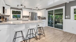 Une cuisine moderne avec des meubles blancs, un plan de travail en granit et un réfrigérateur en acier inoxydable. On y trouve trois tabourets de bar et une grande fenêtre donnant sur un jardin.