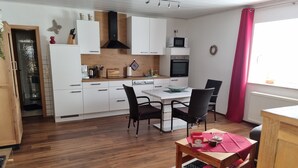 Une cuisine moderne avec des armoires blanches, une hotte noire et un four à micro-ondes. On y trouve un coin repas avec une table et des chaises, un tapis et une fenêtre ornée d’un rideau rouge.