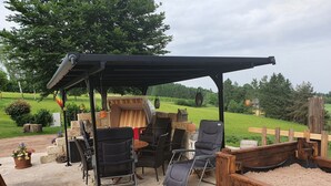 Une terrasse extérieure dotée d’un espace salon couvert, comprenant des chaises et une table, entourée d’une clôture en bois et offrant une vue sur un paysage verdoyant.