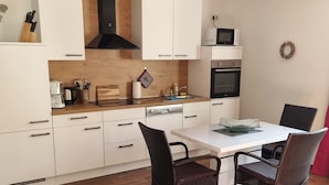 Une cuisine moderne avec des meubles blancs, un plan de travail en bois, un four encastré et un coin repas avec des chaises en osier.
