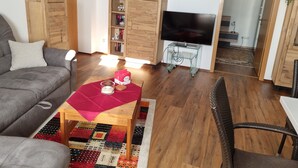 Un salon avec un canapé gris, une table basse en bois recouverte d’un tissu rouge, une bibliothèque, un téléviseur posé sur un meuble et un fauteuil en osier.