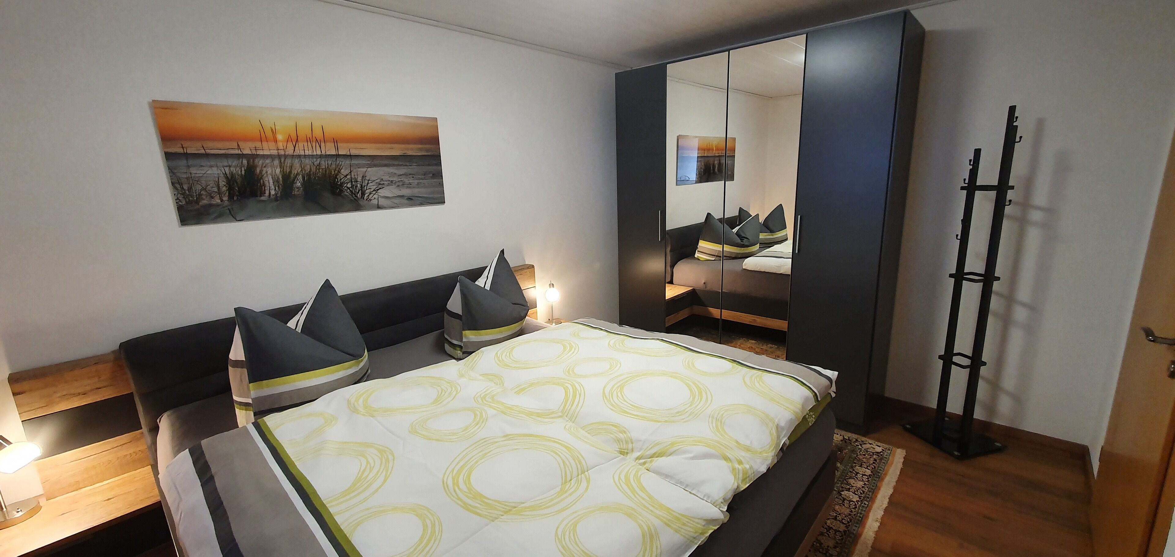 A bedroom with a bed, bedside tables, a mirror, and a painting of a sunset.