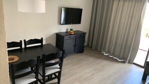 Een eetgedeelte met een tafel en stoelen, een aan de muur bevestigde televisie en een kastje met een fruitschaal erbovenop.