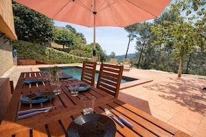 Une table en bois, dressée avec des assiettes, des verres et des couverts, un parasol et une piscine en arrière-plan.