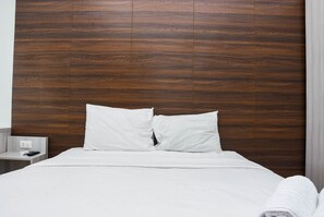 Cama bem arrumada com lençóis e almofadas brancas, cabeceira de madeira e uma pequena mesa de cabeceira com controle remoto.