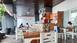 Un restaurant avec des tables et des chaises en bois, une grande plante en pot et un espace cuisine doté de meubles en bois.