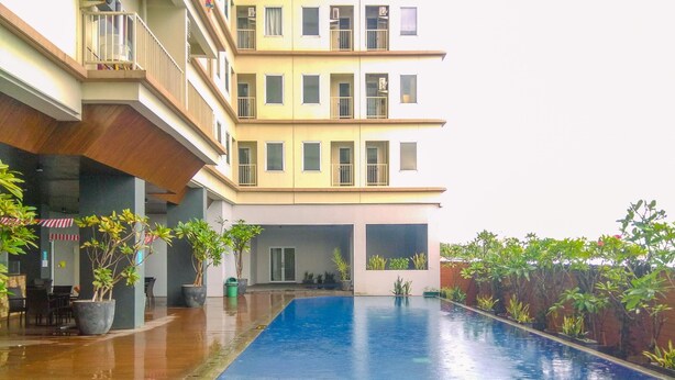 Une piscine dotée d’une terrasse carrelée, entourée de plantes en pot et bordée d’un immeuble à plusieurs étages avec des balcons.