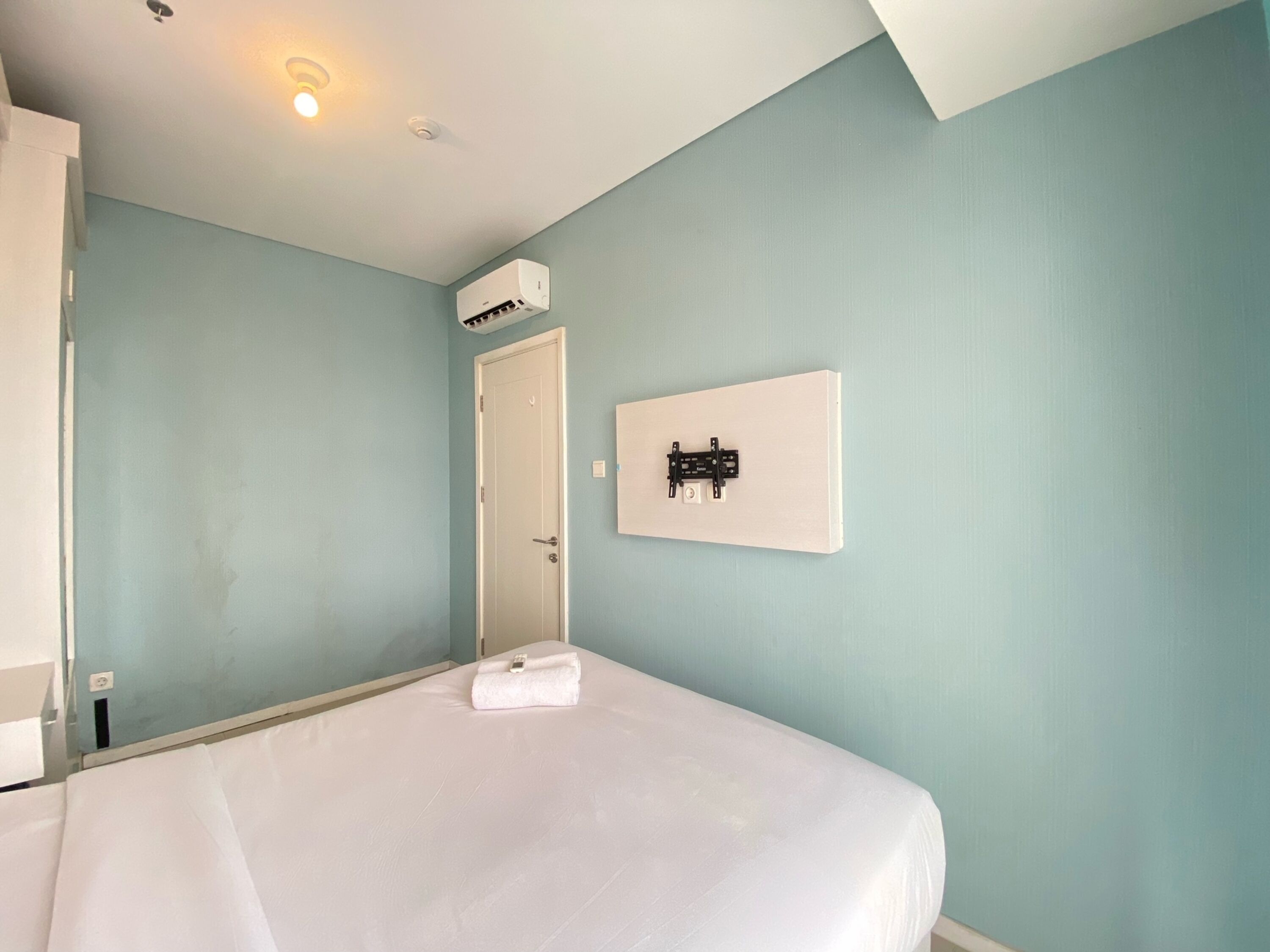 A hotel room with a bed, a wall-mounted air conditioner, a door, and a framed picture on the wall.